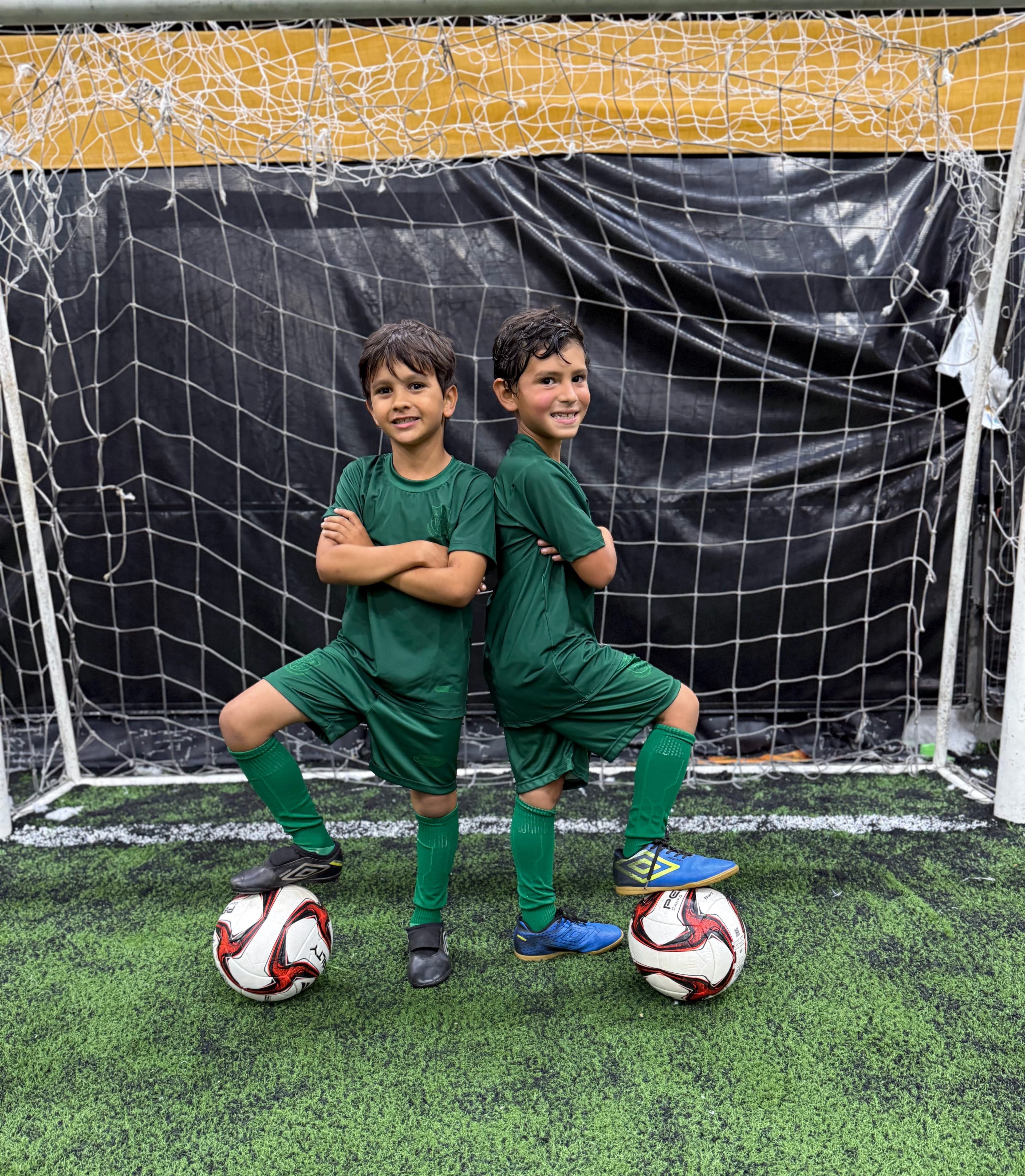 Imagem de duas crianças lado a lado segurando bolas de futebol nos pés. Alunos da Escola Cristã do Esporte, localizada no Raboni Complexo Esportivo